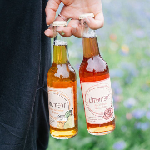 Eine Person hält neben ihren Beinen eine Flasche Limoment Birne-Grüntee natürlicher Eistee und Limoment Apfel-Rose Naturlimo in der Hand.