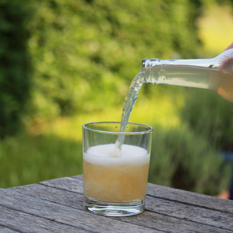 Jemand schüttet eine Limoment naturtrübe Apfel-Spendenschorle in ein Glas, welches auf einem Holztisch steht.
