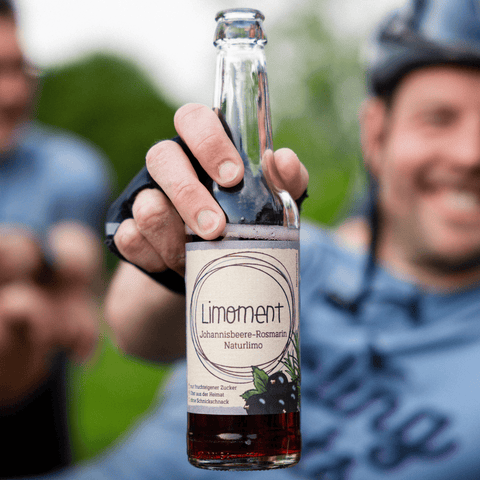 Ein Rennradfahrer hält eine Flasche Limoment Johannisbeere-Rosmarin in die Kamera.