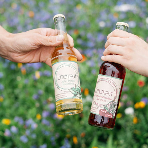 Eine Flasche Limoment Apfel-Minze Naturlimo und Limoment Kirsche-Waldmeister Naturlimo werden vor einer Blühwiese in die Kamera gehalten.