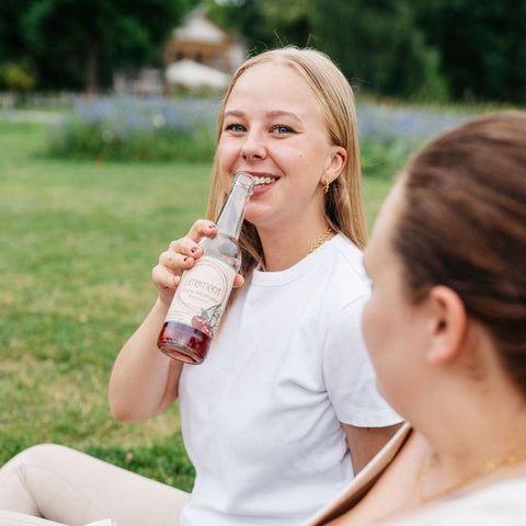 Eine Frau sitzt auf einer Wiese, trinkt eine Flasche Limoment Kirsche-Waldmeister Naturlimo und schaut dabei in die Kamera.