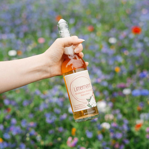 Jemand hält eine offene, sprudelnde Flasche Limoment Rhabarber-Holunder Naturlimo vor einer Blühwiese in der Hand.
