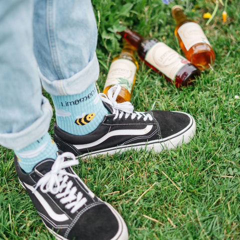 Eine Person mit schwarzen Stuhen steht auf einer Wiese, trägt die Summsocken von Limoment. Hinter ihr liegen Flaschen von Limoment Naturlimos.