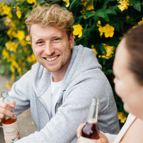 Ein Mann lächelt und hält dabei eine Flasche Limoment in der Hand. Im Vordergrund ist eine Frau zu sehen, ebenfalls mit einer Flasche Limoment.