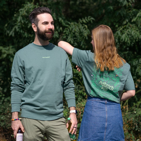 Präsentation des salbeigrünen "Natürlich süß" Pullovers (Vorderseite am Mann) und des T-Shirts (Rückseite mit floralem Print an der Frau) im Grünen.