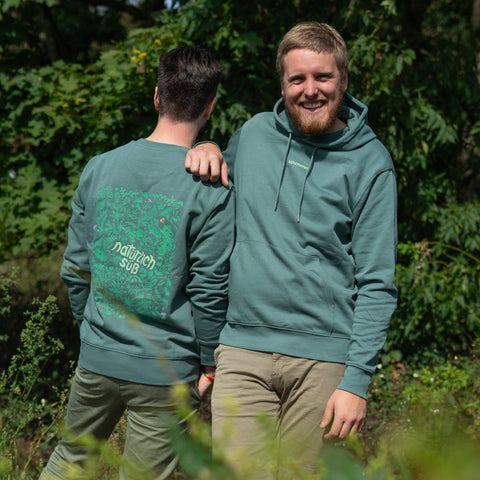 Zwei Männer präsentieren den "Natürlich süß"‘ Merch in Salbeigrün. Der rechte Mann zeigt die schlichte Vorderseite des Hoodies mit Kapuze. Der linke Mann, von hinten gesehen, zeigt die Rückseite des Pullovers (Sweater) mit dem auffälligen, detaillierten floralen Rücken-Print.